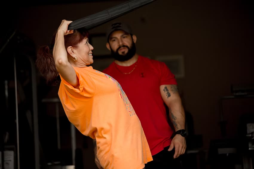 An older woman pressing a weight overhead while Martin coaches beside her
